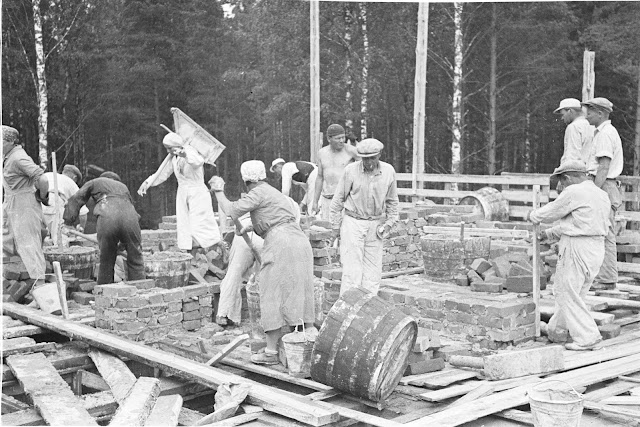 Mustavalkoinen kuva. Miehiä ja naisia rakennustyömaalla. Naiset pukeutuneina hameisiin ja esiliinoihin. Miehillä housut ja paita päällä.