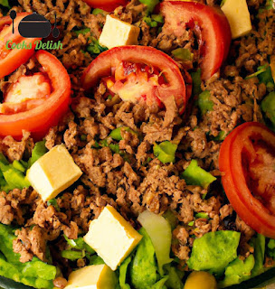 This image shows a colorful bowl of taco salad with lettuce, tomatoes, cheese, olives, and green onions. The ground beef is visible on top, sprinkled with taco seasoning. The dish is garnished with sour cream, salsa, and crushed tortilla chips.