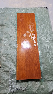 A photograph of a wooden plank on a green tarp. The wood has a resin coating on it that has an oily film in one area, and has separated from the wood in another area.