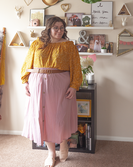 An outfit consisting of a yellow long sleeve microfloral print off the shoulder blouse tucked into a muted pink button down midi skirt and pink slide sandals.