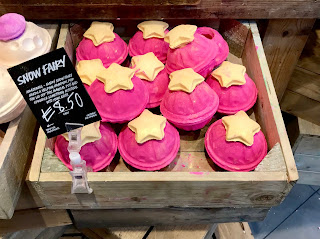A spherical large hollow pink bath bomb with a bright yellow star stopper on top in a large rectangular light brown box on a bright background