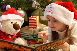 クリスマスツリーの前に腰掛けているサンタの帽子を被った少女とぬいぐるみの白クマ。少女はしろくまに絵本を読み聞かせている。