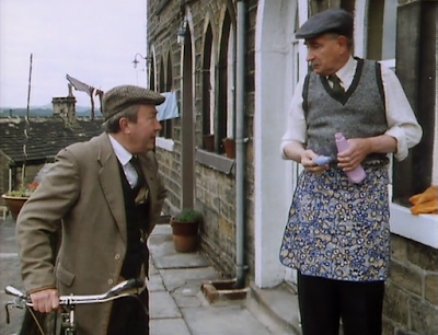 Howard from last of the summer wine cleaning windows with a pullover over his pinny, while Normal Clegg leans on his bike to talk to him