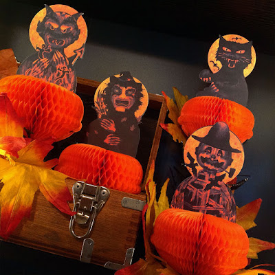 Four characters - black cat, scarecrow, witch, and devil with honeycomb tissue paper on the shelf with leaves and an old crate.