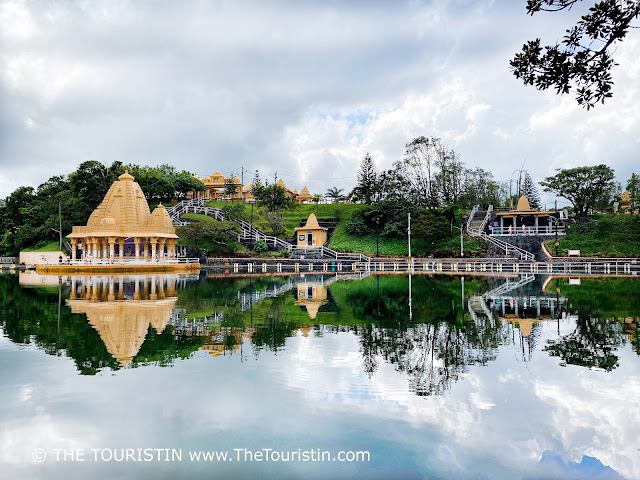 Three midsize yellow-beige temples surrounded by lush vegetation on the edge of a large water basin, all is reflected in the water.