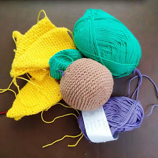 A group of small crocheted items in yellow, brown and green and two balls of cotton yarn - one green and one purple.  It isn't clear what the objects are.