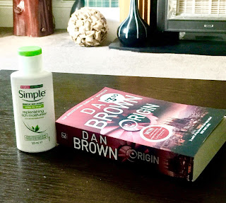 A tall rectangular white bottle with a green cylindrical lid that says simple in big green font then Kind to skin moisturising cream in small black font next to a rectangular book saying dan brown in white font on a brown rectangular table  on a bright background 