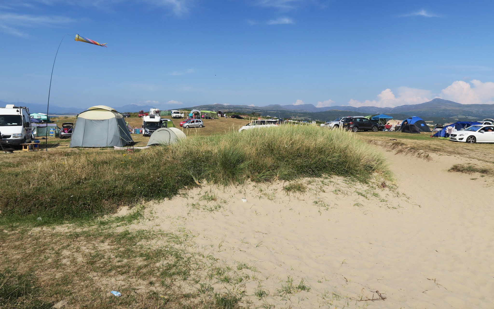 Standing on a sandy path looking towards the campsite. Tents and campervans can be seen across the landscape.