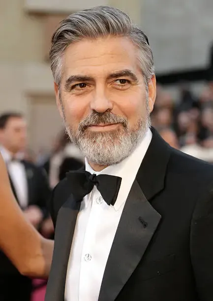 George Clooney é uma das maiores estrelas de Hollywood, conhecido por seu charme e beleza icônica. Clooney tem um rosto magnífico, com uma testa alta e olhos escruos profundos, cabelo grisalho, queimado ao sol, e uma barba desalinhada, que lhe dão um ar misterioso. Seu charme é irresistível, e seu sorriso cativante.