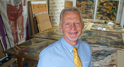 image of an old white man smiling in front of a large table that has paintings on it