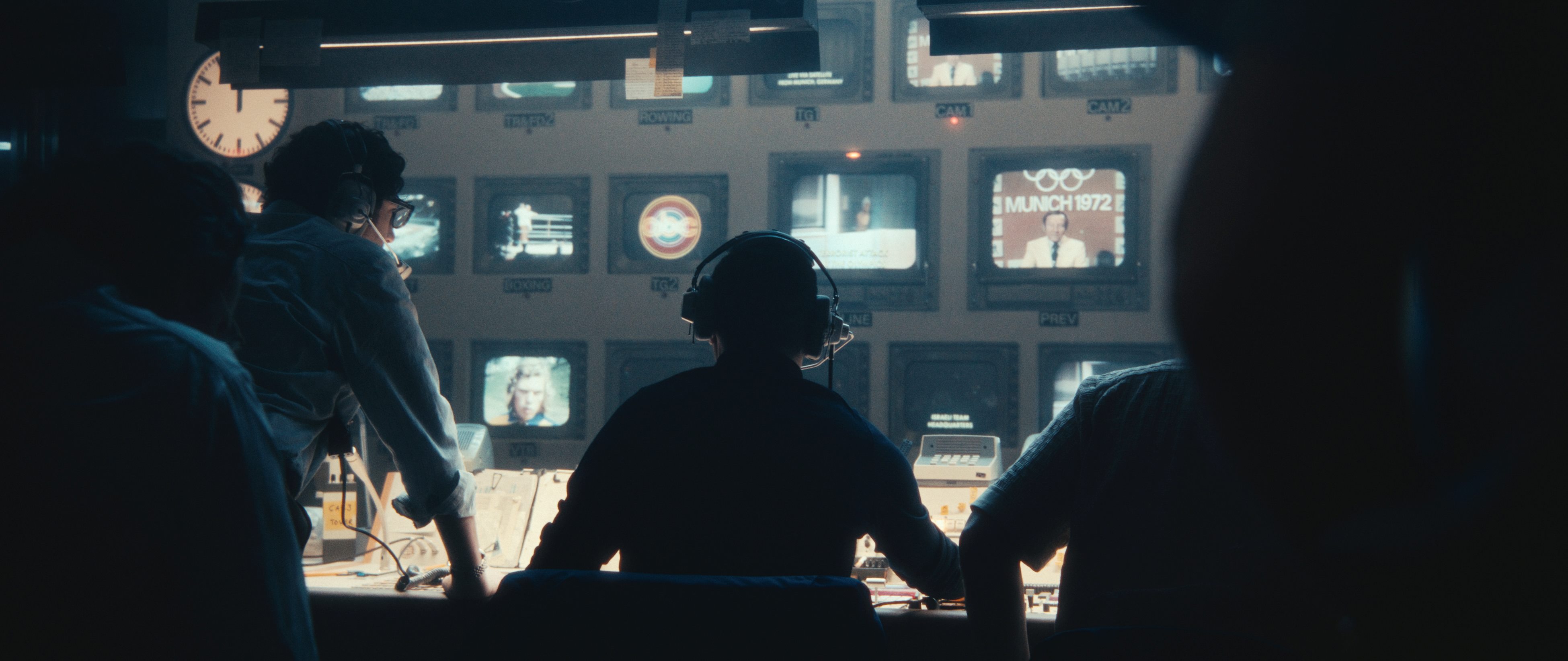 Several men are positioned facing a brightly-lit control panel and several televisions in an otherwise dark room.