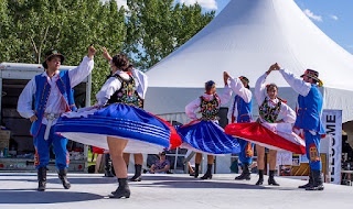 Color photo of men and women in traditional costume folk-dancing. The men's clothes are blue breeches and jackets with red accents, a white shirt, and black hat and boots. The women are wearing full blue or red skirts, white aprons, black bodices with colorful embroidery, white blouses, and black boots. The womens' hair is braided with red bows and red or blue flowers.