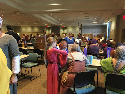 A large room with rows of tables and green-cushioned office chairs, with several people in bright medieval garb standing and sitting around them. A young girl with long blond hair and a red bliaut is leaning on the shoulder of a grey-haired woman in white shift and madder-pink apron dress in the foreground..