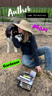 dry grass covered garden surrounded by a black fence and in the middle sits a blonde woman wearing  a large hat, blue jeans, and black hoodie, as she hugs her black and white Siberian husky