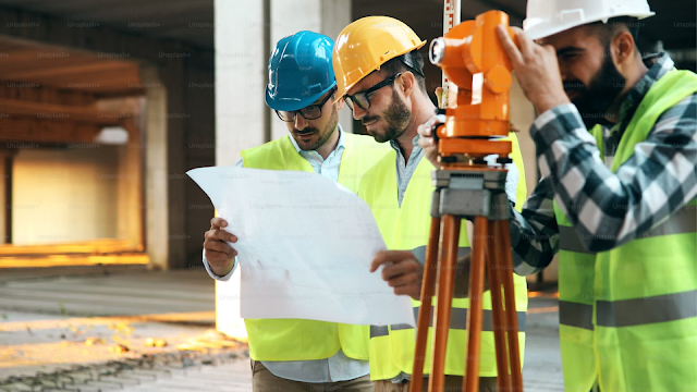 Professionals in safety helmets and reflective vests examining a blueprint beside a theodolite tripod instrument at a construction site.