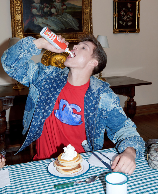 Photo of male model sitting at breakfast tabel wearing preloved designer clothing. Model wears red Dolce and Gabanna T-shirt with purple branding and a Patchwork Louis Vuitton Denim Jacket