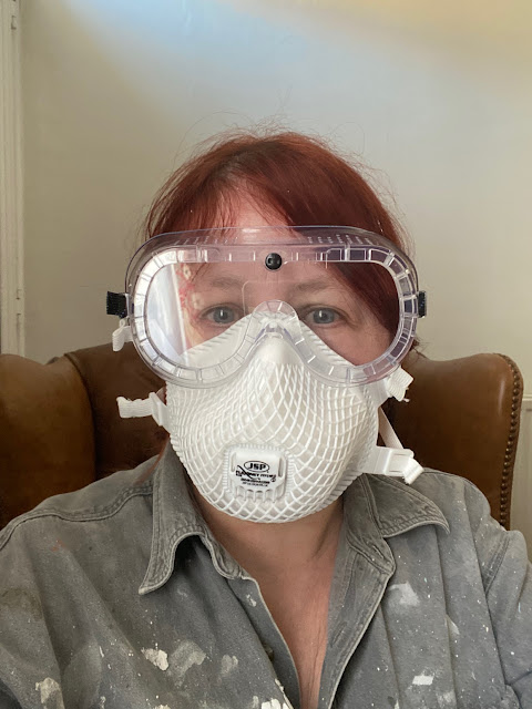 A red-haired white woman wearing goggles, a breathing mask and a paint-spattered grey denim shirt