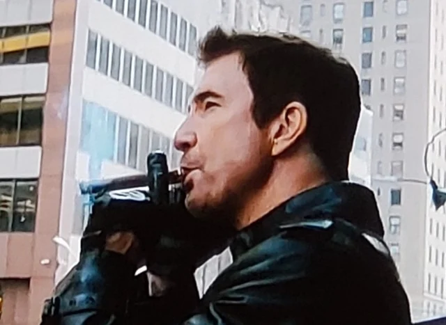 Dylan McDermott wearing a black leather jacket and gloves and smoking a cigar from the chest up in a city