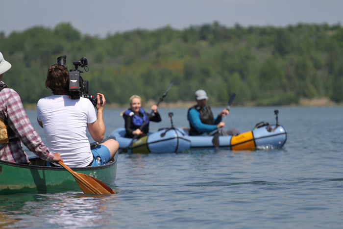 Auf dem Bild sind drei Boote: Zwei Packrafts im Hintergrund und ein Kanu mit einem Kameramann im Vordergrund. Der Kameramann filmt die Packrafts während des Paddelns.