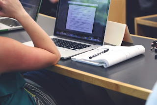 A picture zoomed in on two pale hands on a black rectangular keyboard below a large swuare black and glass computer monitor with a white mouse at the side of it on a rectangular white table on a bright background. 