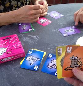 A photo of the hands of three people playing Simon's Cat. The cards in their hands, and on the table, are coloured according to the animal on the card (a cat, a mouse, a kitten, and so forth). Each card is numbered.