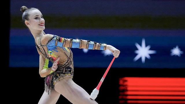 Darja está de collant colorido segurando as maças e sorrindo. Ela é morena e branca