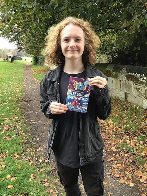Will stands with windswept blond hair outside on a pathway in a park wearing a black tshirt, an open black denim jacket and black trousers holding a book called The Rescue of Ravenwood up.