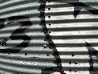 A picture of the corrugated surface of one of the metal containers.  On one of a set of four rusty screws on the container sits the small, ceramic skull (Skulferatu 119).  Photo by Kevin Nosferatu for the Skulferatu Project.