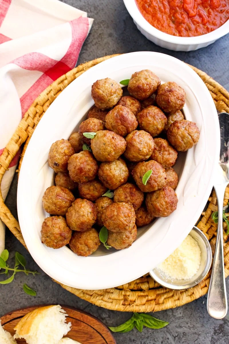 Air-fried meatballs in a white oval bowl sitting on a wicker plate holder with a small silver bowl of parmesan cheese and a red and white checkered napkin sitting next to it.