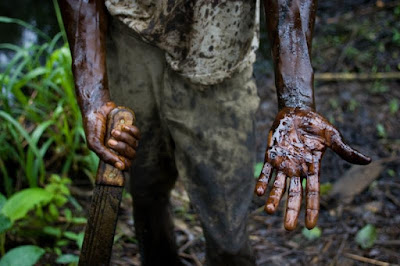 Worker cleaning oil spil inNiger Deltal holds out his oils-covered arms and hand with a machette in one hand