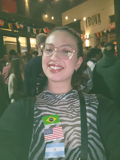 Selfie de uma mulher em um bar decorado com bandeirinhas de países. Ao fundo lê-se Trova, o nome do bar. Na vestimenta da mulher estão coladas três bandeirinhas, a do Brasil, a dos Estados Unidos e a da Argentina, respectivamente. É uma selfie do evento Mundolingo.