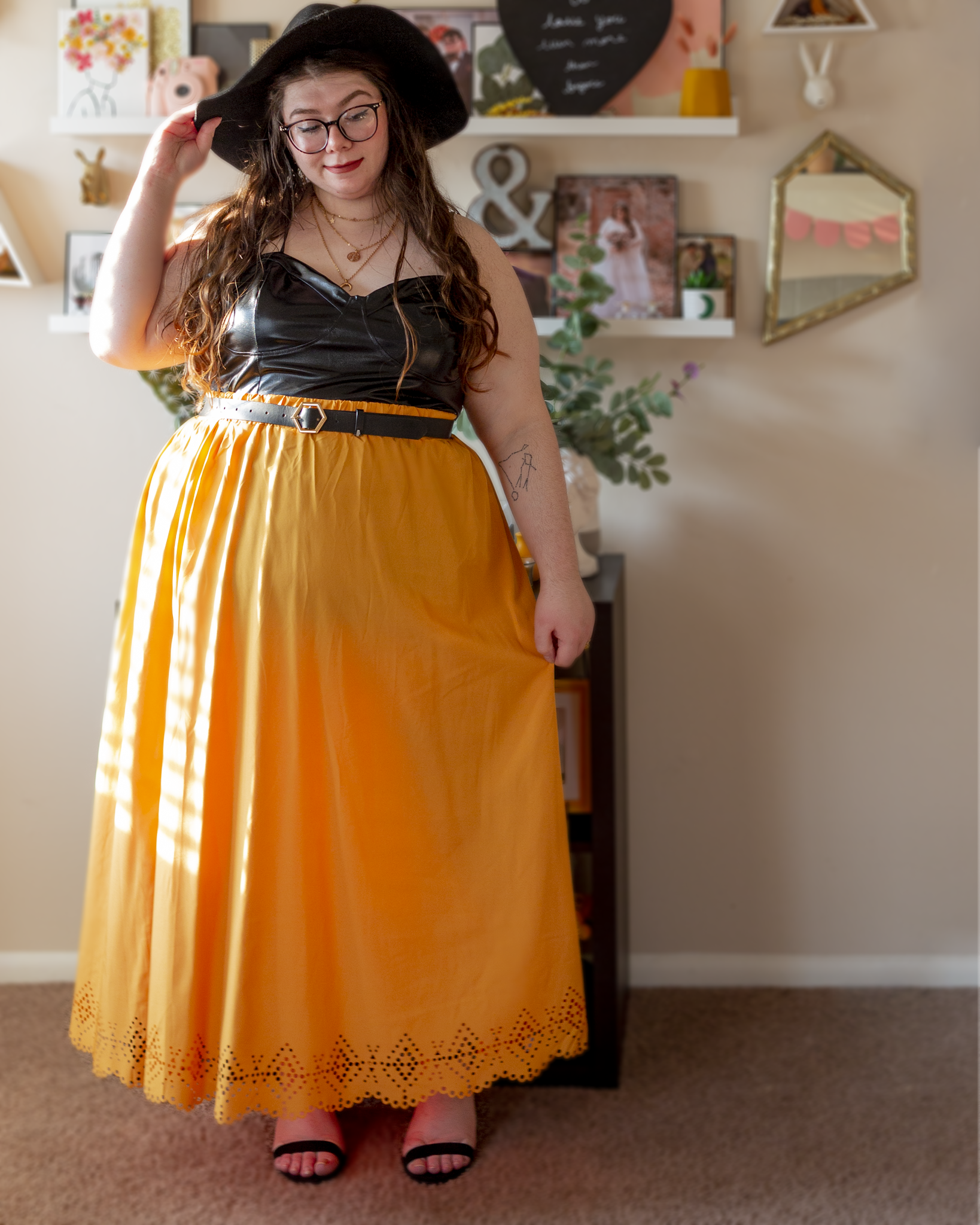 An outfit consisting of a black wide flippy brim fedora, a black vinyl camisole crop top tucked into a mustard yellow scalloped laser cut hem maxi skirt, belted with a black belt and black ankle strap heeled sandals.