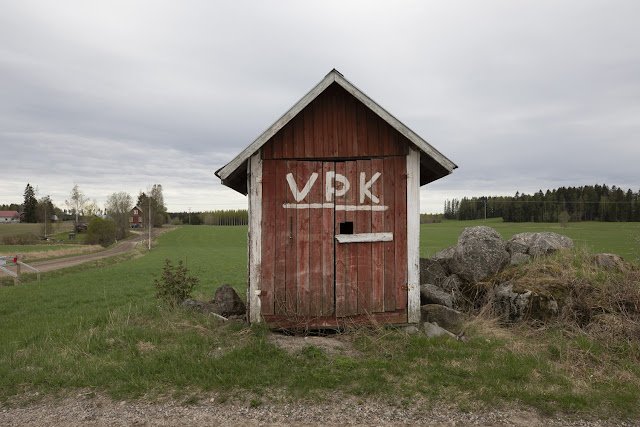 Pellon reunassa oleva pieni punainen lautarakennus, jonka ovessa lukee VPK.