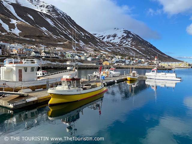 White and yellow fishing boats and their reflections in the calm water of a fjord in front of a snow-capped mountain range under a blue sky.