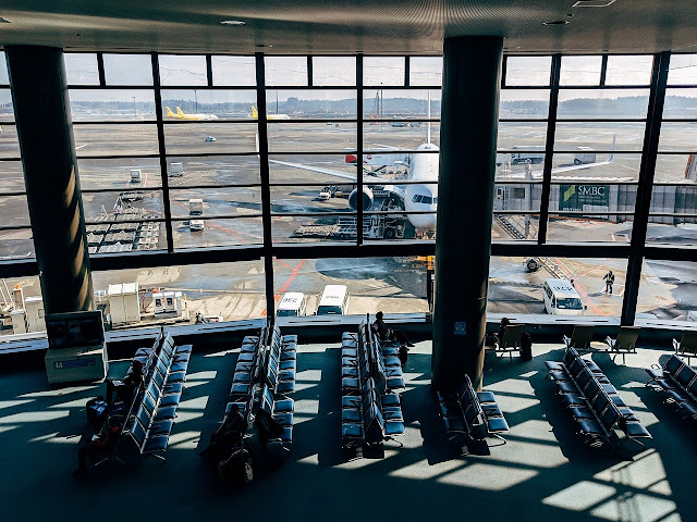 Looking down on an airport gate area. Bright sunlight streams in. Tarmac is visible outside, with airplane docked to the gangway.