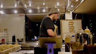 A tall pale man wearing a grey beanie hat, dark blue shirt, black jeans and a white apron standing over a silver large square metal over in a white rectangular van on a bright background 