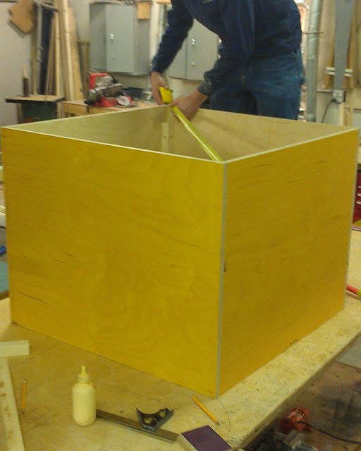 On a table sits the casework.  It is a yellow box with no top that measures 34" by 34" by 34".  A young man in a long sleeved dark blue shirt stands behind it on the table measuring the diagonal distance between two points on the top of the box with a yellow tape measure.  A bottle of yellow colored wood glue can be seen in front of the box and the grey cabinets can be seen on the wall in the background.