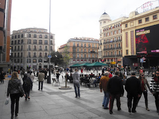 Numerosas personas transitan por esta plaza, mientras otras descansan sentados en una terraza.