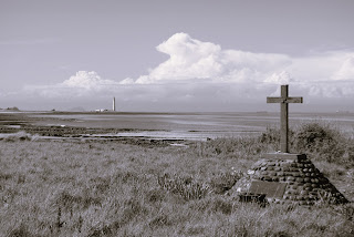 A black and white photo showing a memorial cross sitting on a triangular pedestal lined with pebbles.  In the distance Barns Ness Lighthouse can be seen.  Photo by Kevin Nosferatu for the Skulferatu Project.