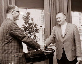 Tom Roos and President Kemeny shake hands in front of a small table with a potted lemon tree.