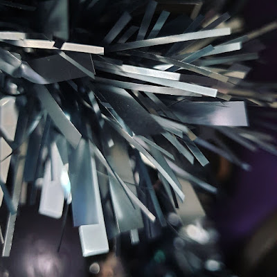 Grey tinsel above a blurred purple bauble and against the background of a dark purple wall. The fronds of the tinsel are quite chunky, the tinsel is dark grey and metallic