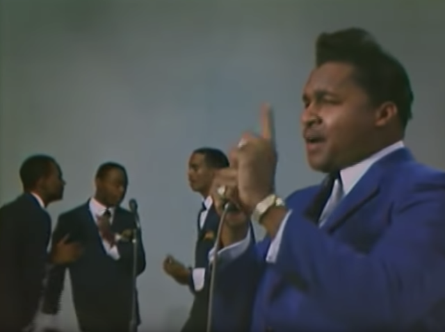 photo Little Milton performing on The Beat tv show in 1966 wearing a blue suit, holding a microphone in one hand, holding up his index finger with the other hand, and with three back up singers in the background.