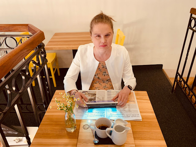 Linda sedí v kavárně u stolku, pomocí přenosné lupy čte text v časopisu. Na stole je hrnek s kávou, konvička s horkou vodou a konvička s mlékem. a květina ve váze.