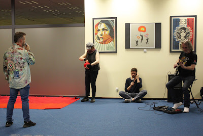 Na zdjęciu osoba zwija kłębek czerwonej wełny, ten proces  jest część performancu, obok osoby grają na instrumentach, prowadzący mówi przez mikrofon