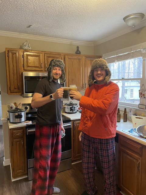 Dad and Andy standing in the kitchen with their Russian hats on. Andy has on his orange coat and dad is in a tshit. They each have a beer mug in their hands and are clinking their glasses together.