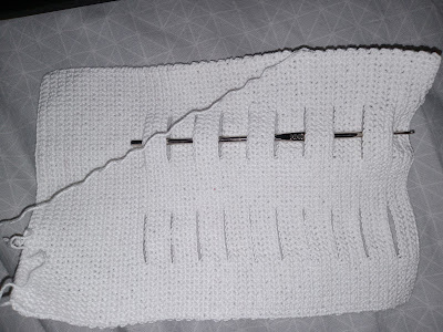 A rectangle of white crocheted fabric with the leading thread stretched across at a diagonal from the upper right corner to the middle of the left end. The piece is on its side. There is a double-line of slits starting an inch and a half from the left side and leading to the right side. The steel hook is woven through the top row of slits.