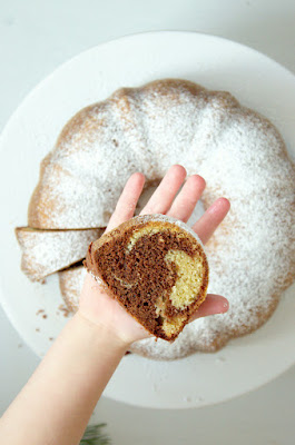 Auf einer Kinderhand liegt ein Stück Marmorkuchen. Darunter ist unscharf der ganze Kuchen zu erkennen.