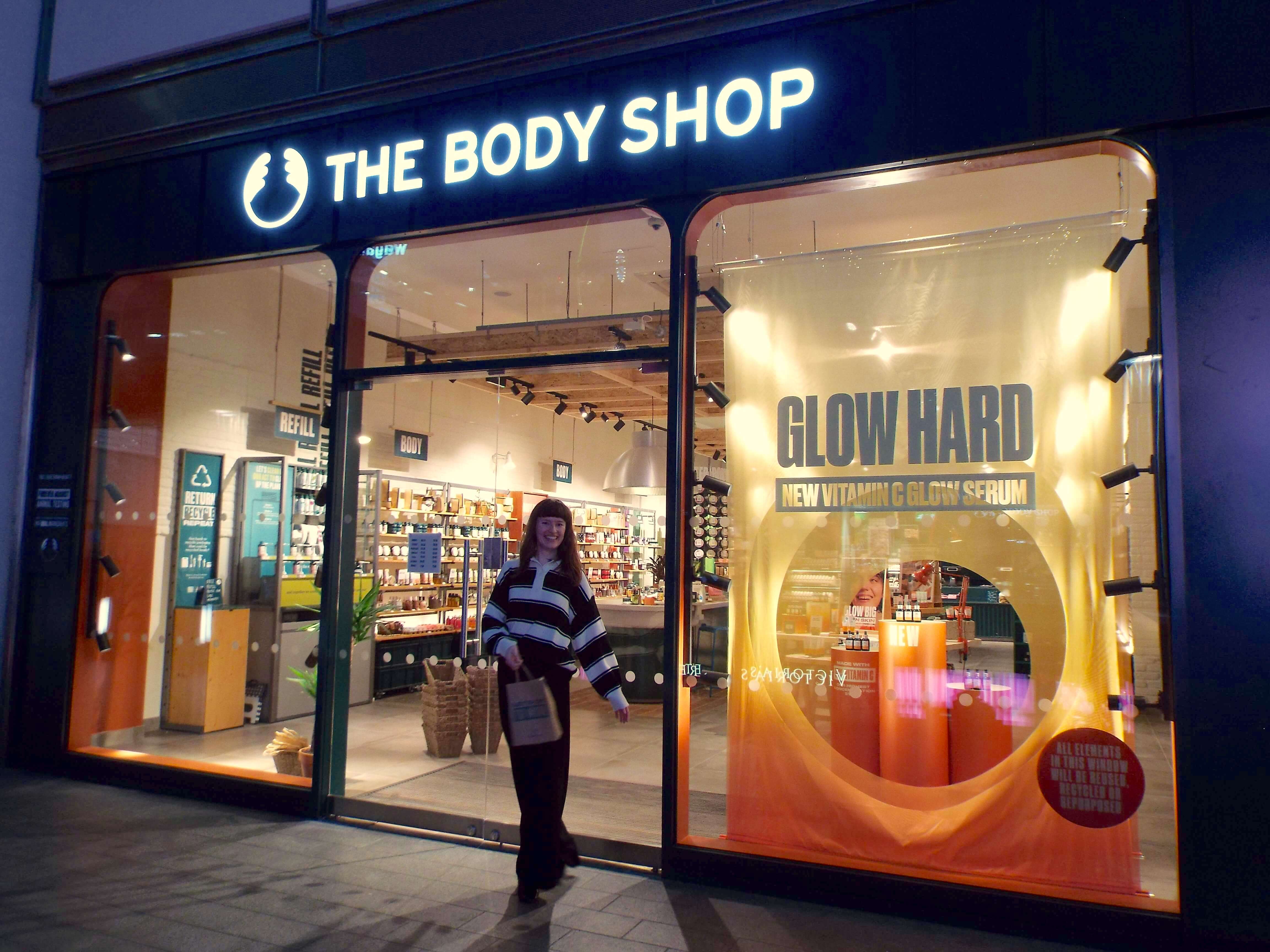 Ellie, cheerfully posed, outside The Body Shop Liverpool One, with a warm exterior to the shop front.