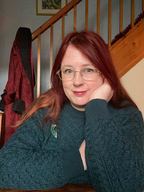 A redheaded white woman in a chunky Aran sweater. It's a deep teal colour, with heavy cable and honeycomb stitch patterns.