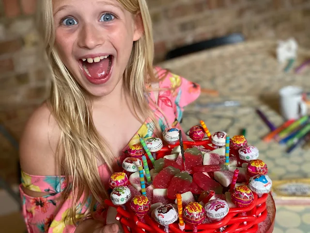 An excited birthday girl at her party holding her birthday cake which is completely made from sweets from a liquorice and lollipop outside to lots of soft jelly sweet fillings